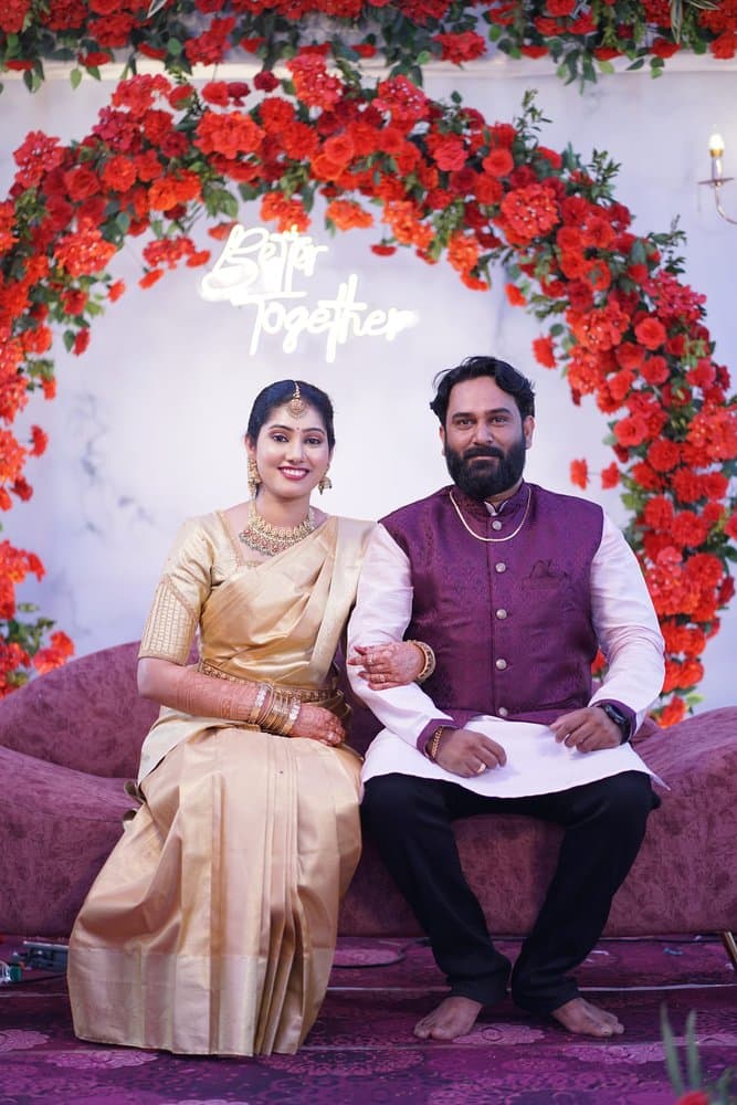 Shravya and Pramod sitting side by side on a burgundy sofa under an arch of orange roses, holding hands