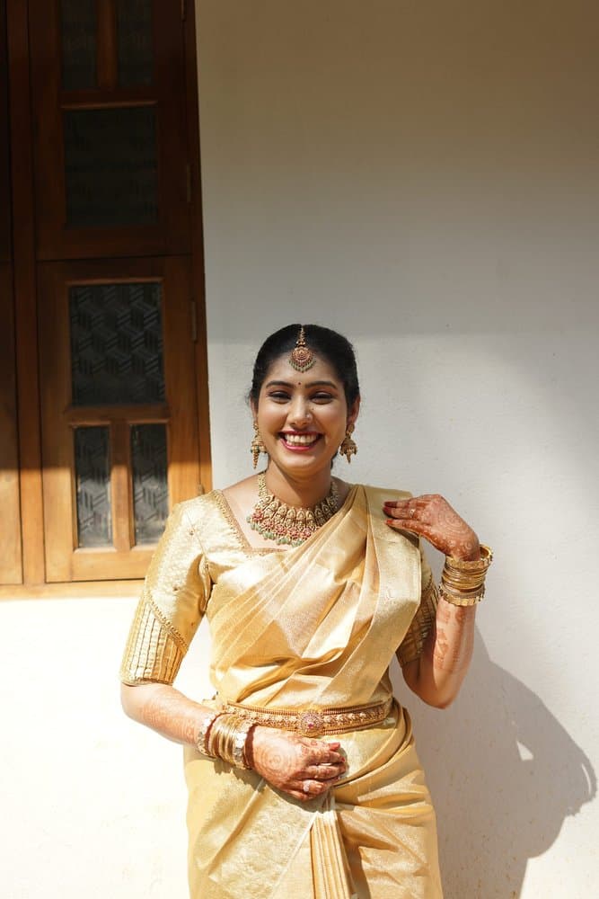 Shravya laughing in a gold silk saree and temple jewellery, standing against a sunlit white wall