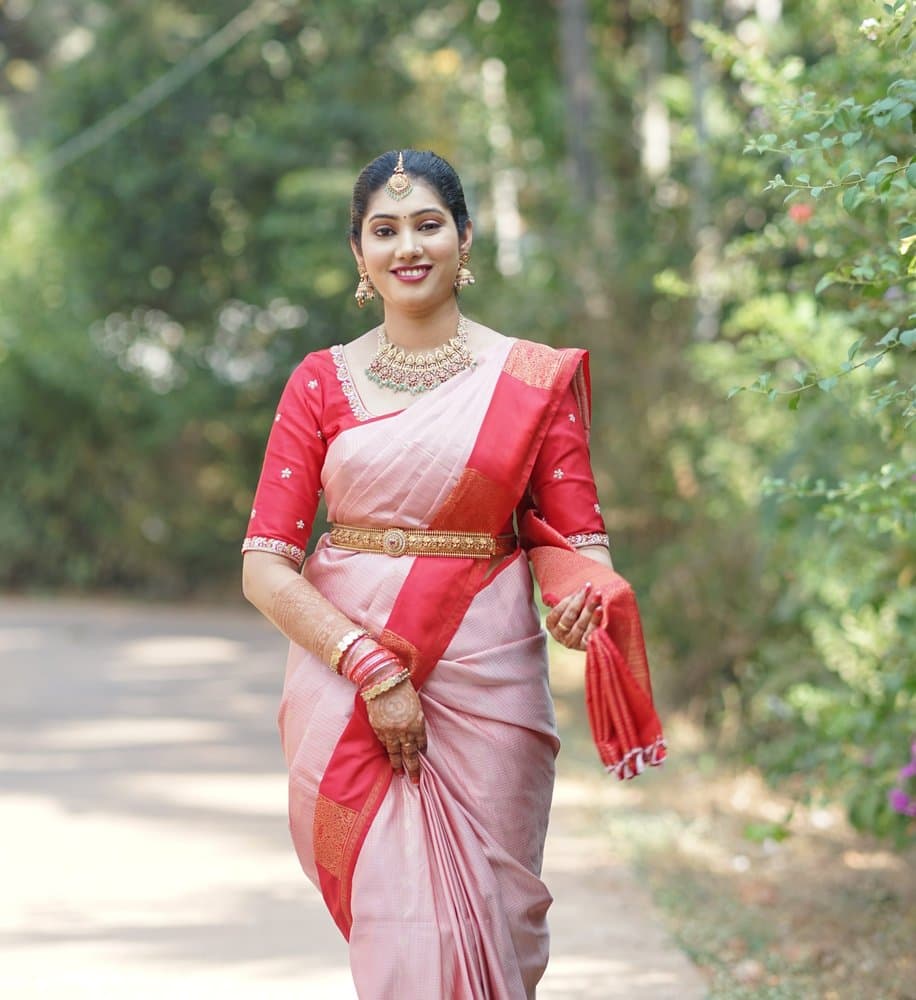 Shravya smiling as she walks along a tree-lined path in a pink and red silk saree