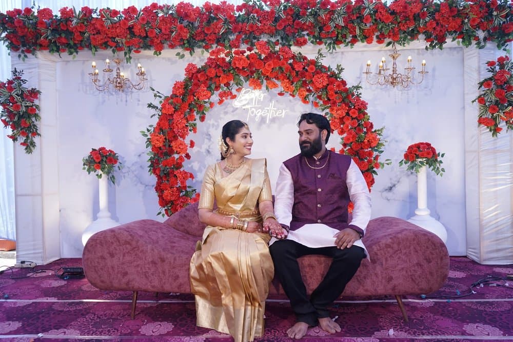 Shravya smiling at Pramod as they sit hand in hand on a burgundy sofa under an arch of orange roses