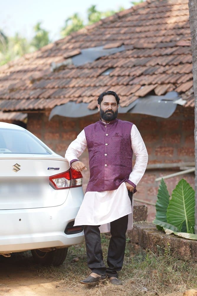 Pramod smiling in a purple brocade jacket over a white kurta, standing in front of a tiled-roof house